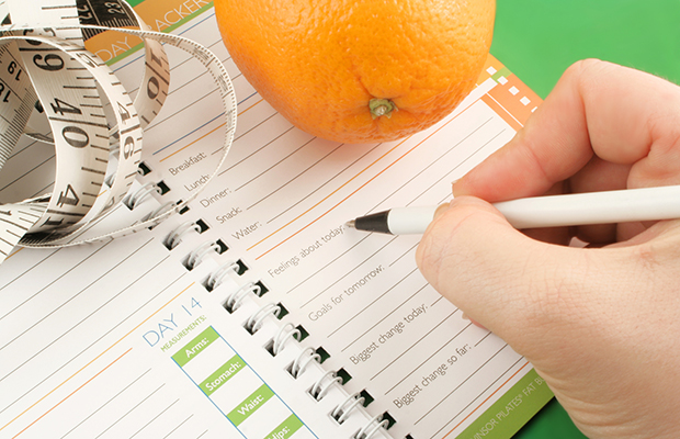 writing in a diet and nutrition journal with orange and tape measure to the side